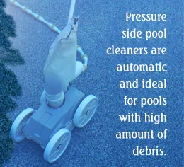 A blue automatic pool cleaner with wheels sits on a blue textured surface next to a green bush.