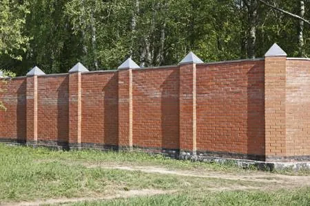 A long red brick fence with decorative pyramidal caps stands in a grassy area backed by a line of trees.