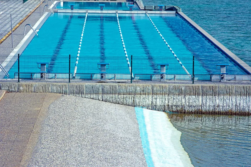 Blue swimming pool with lane markers and starting blocks enclosed by a black metal fence next to the ocean.