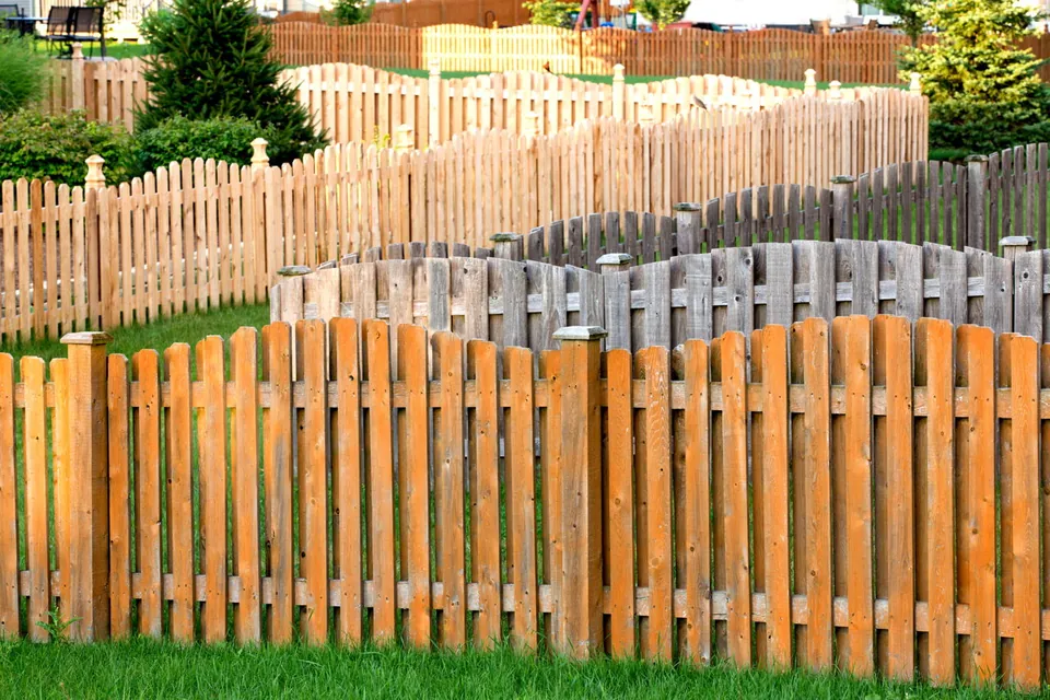 A display of wooden picket fences in varying shades of natural, grey, and stained wood curves across a grassy lawn.