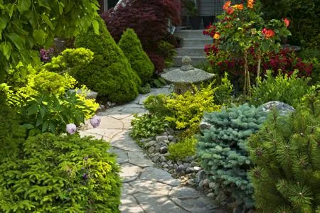 A stone pathway winds through a lush garden filled with green shrubs, a stone lantern, and steps leading upward.