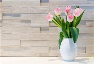 Pink tulips in a white textured vase on a white surface against a light wood plank wall.