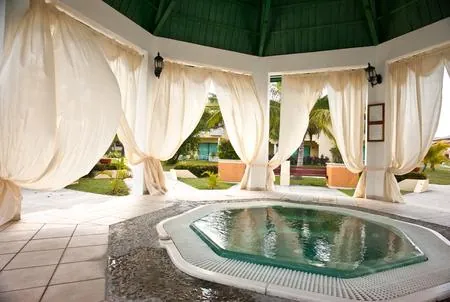 An octagonal hot tub sits under a white gazebo with billowing curtains and a green roof, overlooking a manicured garden.
