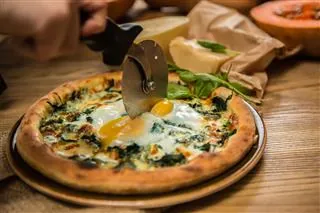 A pizza topped with fresh arugula and eggs is being sliced on a wooden table.