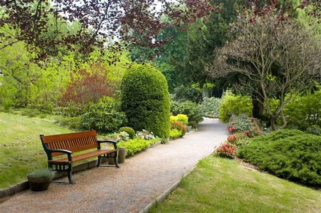 A wooden bench sits along a winding gravel path in a lush garden with manicured shrubs, colorful flowers, and mature trees.