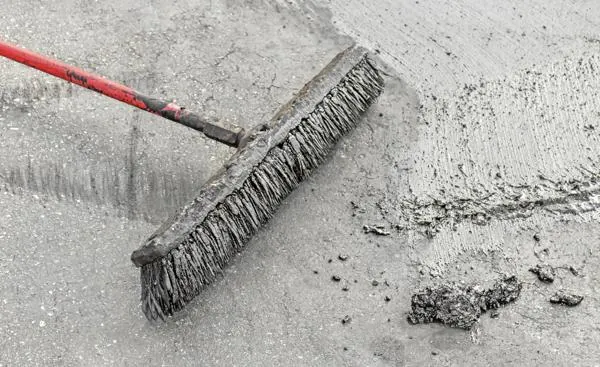 A red-handled broom brushing wet concrete resurfacer on a gray driveway.