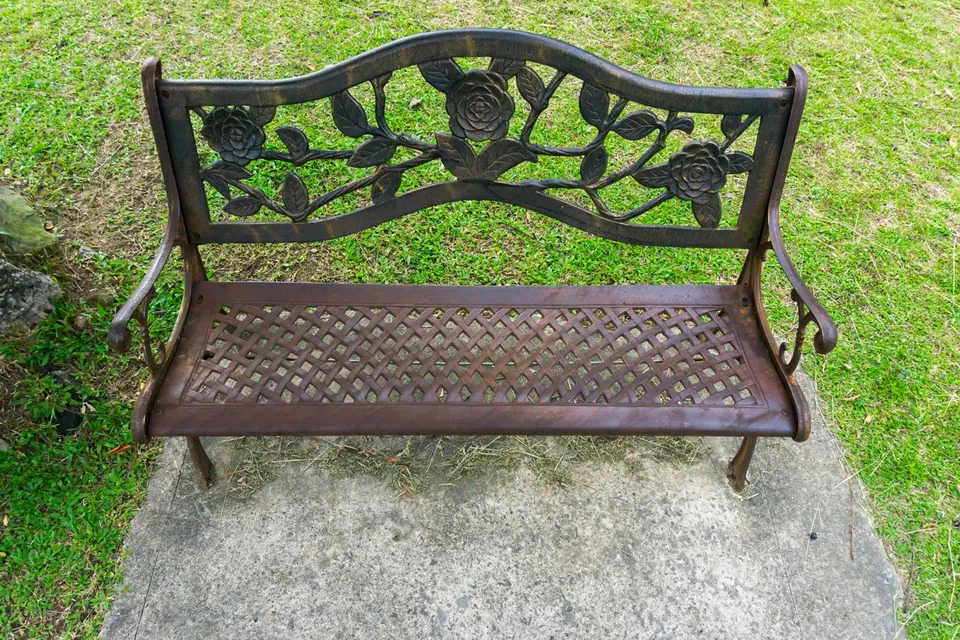 A dark metal garden bench with a rose design on the backrest sits on a concrete slab in a grassy yard.