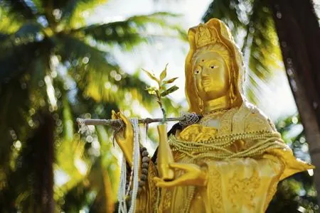 A golden Kuan Yin statue holding a willow branch stands in a garden with palm trees in the background.