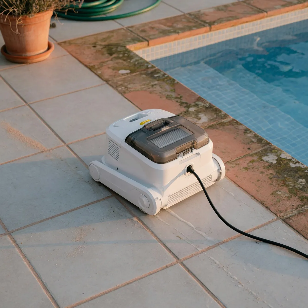 A compact robotic pool cleaner resting on a tiled deck beside clear blue water.